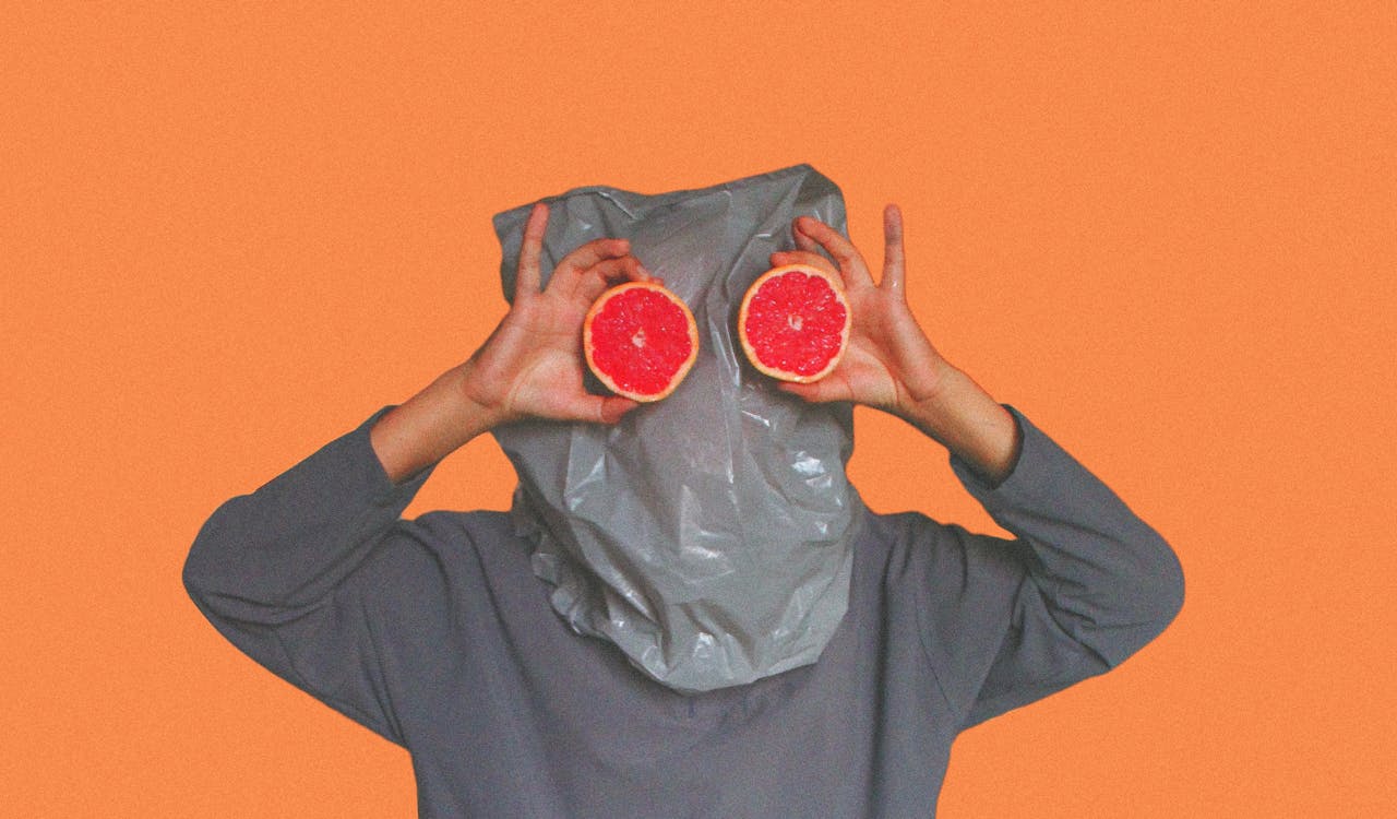 Whimsical portrait featuring grapefruit slices held in front of face with gray bag mask, set against orange background.
