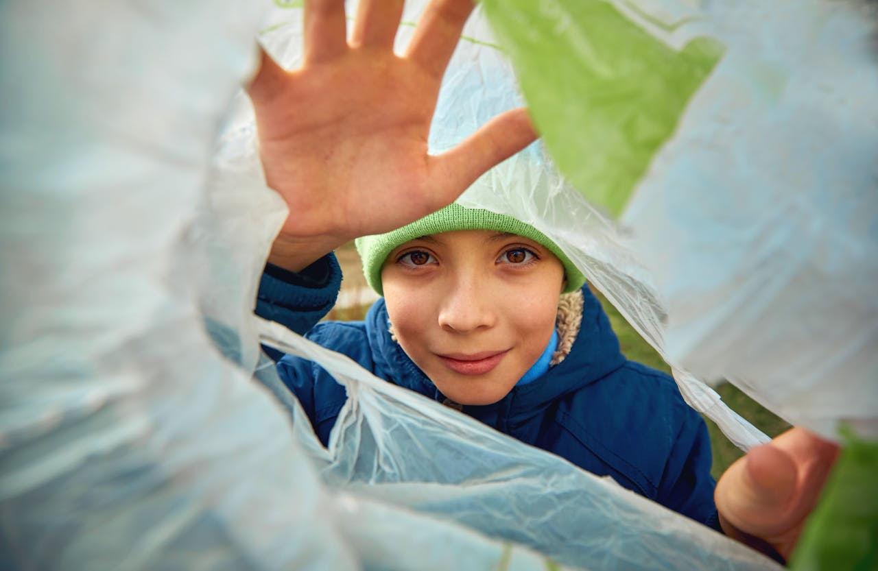 A young child wearing a beanie peers through a recycled plastic bag with a smile.
