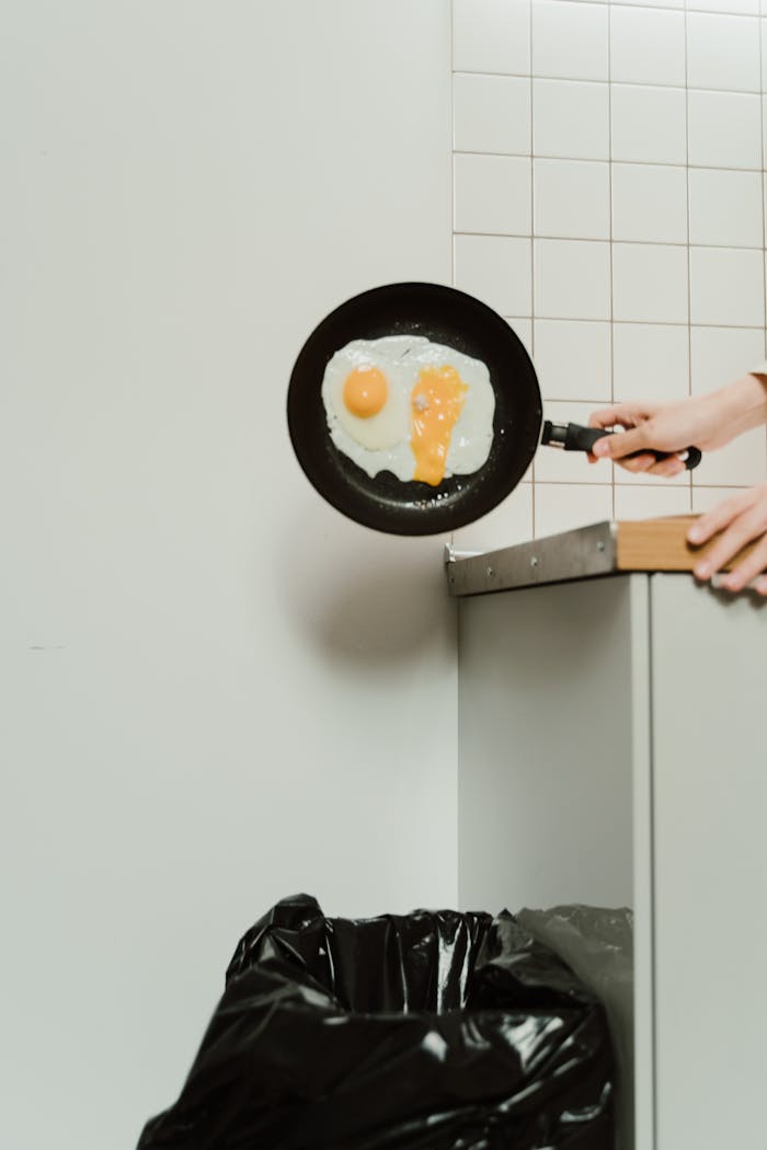 A frying pan with eggs is tossed over a trash can, indicating food waste.
