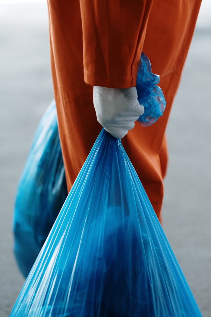 Close-up of a garbage collector in orange uniform holding blue trash bags outdoors.